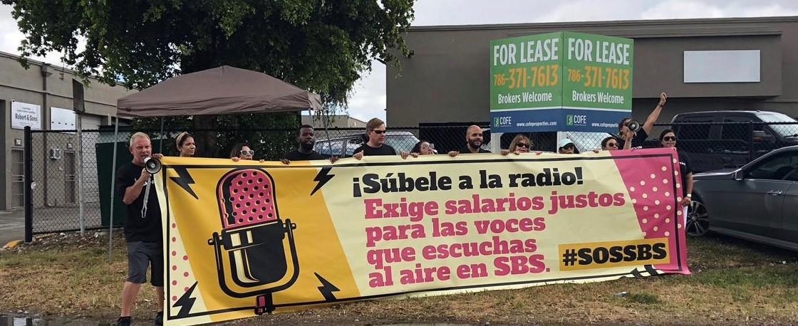 SAG-AFTRA members holding a beige an yellow sign with a pink broadcaster microphone and text to the right that reads "¡Súbele a la radio! Exige salarios justos para las voces que escuchas al aire en SBS. #SOSSBS