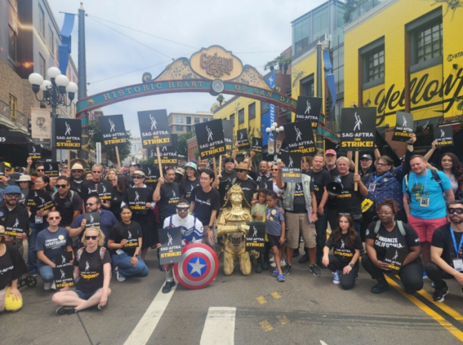 A group photo of a large group in the street. Some in front either kneel or sit on the ground; two individuals are dressed as Captain America and C3P0. “SAG-AFTRA On Strike!” pickets are seen throughout. Also visible is the Gaslamp Quarter arch.