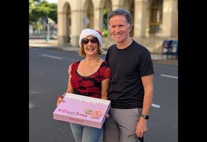 A man and a woman stand on a sidewalk with an older historic building in the background. The woman holds a large pink box of donuts. Both are smiling.