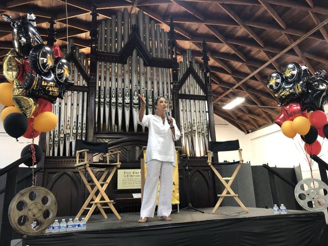 Dodge, dressed all in white, speaks while holding a microphone. She is standing on a stage in front of a pipe organ. There are balloons on either side.