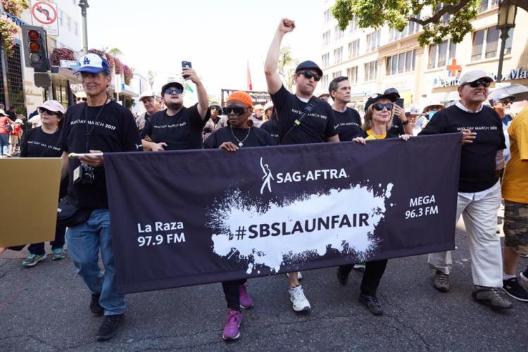 Members wearing Black "May Day March 2017" shirts marching behind a black banner that says "La Raza 97.9 FM" on the left, #SBSLAUNFAIR in black on a white paint splatter design and "MEGA 96.3 FM" in white on the left.