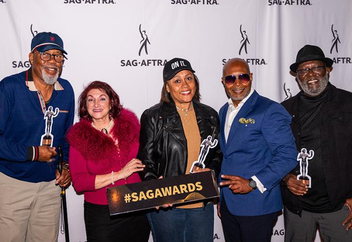 Five member smile as they pose in front of a banner with SAG-AFTRA logos. They are holding replicas of The Actor statuette and a sign that reads "#SAGAWARDS.".