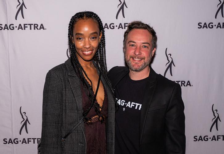 A women and a man smile as they pose in front of a banner with SAG-AFTRA logos.