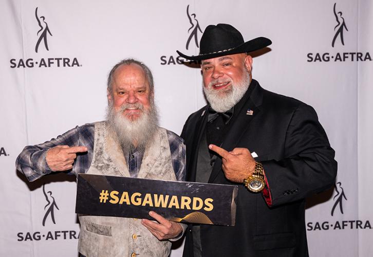 Two men smile as they pose in front of a banner with SAG-AFTRA logos. The man on the left holds a sign that reads "#SAGAWARDS"