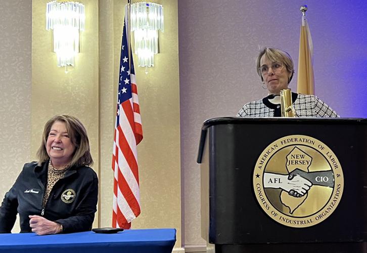 Brennan sits at a table smiling next to an American flag while Pelletier stands at a podium