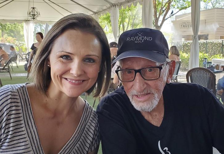 Two individuals are sitting closely together in a relaxed and friendly atmosphere under a tented canopy with smiling faces. The woman on the left is wearing a striped shirt and the man on the right has a white beard and glasses, wearing a SAG-AFTRA T-shirt and a baseball cap.