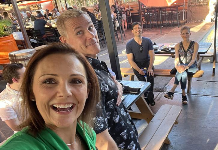 A small group of people enjoy an outdoor gathering at a casual setting with picnic-style benches. The foreground features a woman in a green top smiling brightly and taking a selfie. Beside her is a man in a patterned black shirt, also smiling and looking toward the camera. In the background, a man in a navy shirt sits on a bench near a woman in a patterned top, both smiling and engaged in the scene.