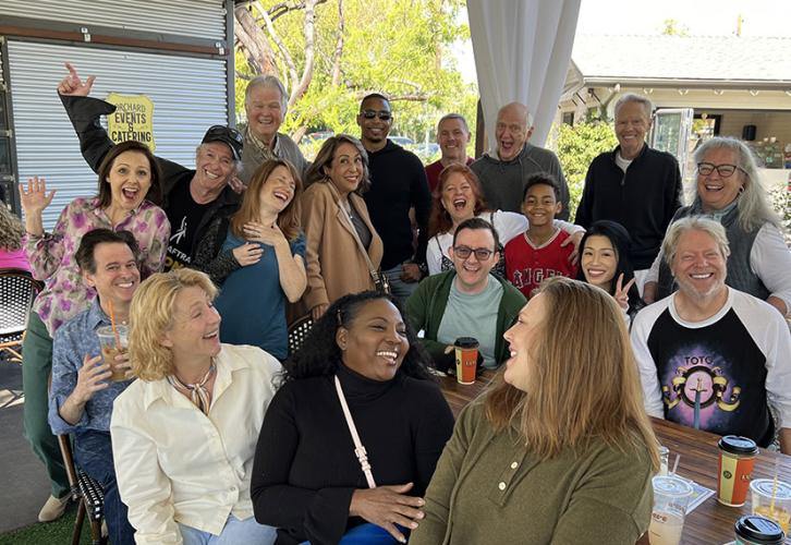 A group of cheerful individuals gather in a sunny outdoor space. The group is diverse in age and style, seated or standing close together around a wooden table with coffee cups and drinks visible.