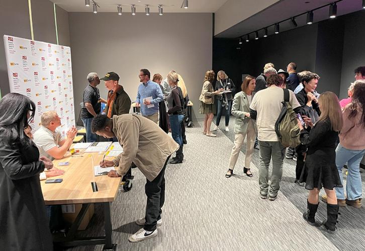  A lively networking event is taking place in a modern indoor setting. Several people are engaged in conversations and enjoying food and beverages. To the left, a wooden table is set up with member volunteers assisting attendees, providing materials and answering questions. Behind the table is a step-and-repeat banner displaying the ASU logo and the name "MIX” Center." Attendees of diverse backgrounds are scattered throughout the room, some chatting in small groups while others browse materials.