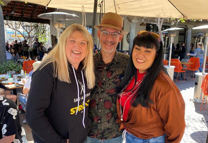 Three people stand outdoors in a lively and sunny setting under a patio umbrella, smiling. The person on the left is a blonde woman wearing a SAG-AFTRA hoodie. In the center, a man wears a patterned shirt, glasses, and a brown hat. On the right, a woman with long dark hair is dressed in a rust-colored top.