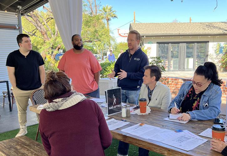 A group of individuals are gathered around an outdoor wooden table under a canopy. Three men are standing on the left, with one man in a navy jacket gesturing as he speaks. On the right, four individuals are seated at the table. The table is covered with papers and a SAG-AFTRA sign.