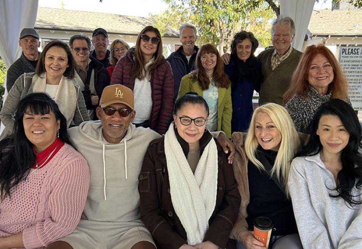 A group of 16 people outdoors during the day posed in two rows, with some sitting in front and others standing behind. They are dressed warmly and appear to be enjoying a casual gathering.