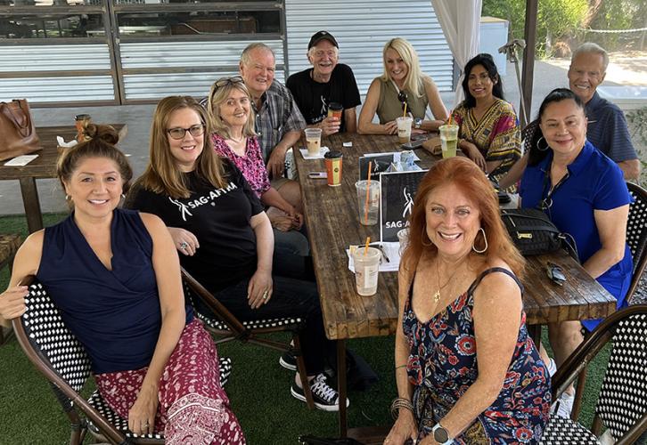 A group of 10 members are seated around a wooden table outdoors. The group is smiling and some individuals are wearing SAG-AFTRA shirts. The table is covered with beverages and a SAG-AFTRA sign.