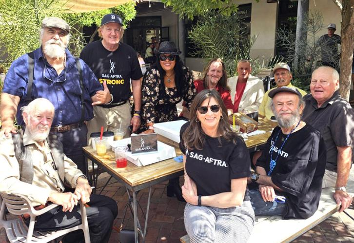 A group of members are gathered outdoors around a wooden table in a sunny atmosphere. Some people are seated, while others are standing behind the table. Several individuals are wearing SAG-AFTRA-branded shirts. On the table are drinks, snacks and a SAG-AFTRA sign. The participants are a mix of men and women representing a range of ages, and are dressed casually, with some in hats and sunglasses.