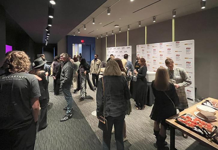 A networking event is taking place in a spacious, modern room. A group of people is mingling and conversing in small clusters, creating a lively and interactive atmosphere. To the right, a table is set up with refreshments. Nearby, attendees are standing by a step-and-repeat banner displaying the ASU logo and "MIX Center." The lighting is soft but focused, with overhead spotlights creating a warm ambiance. The room features dark walls and gray carpeting, giving a sleek, professional vibe.