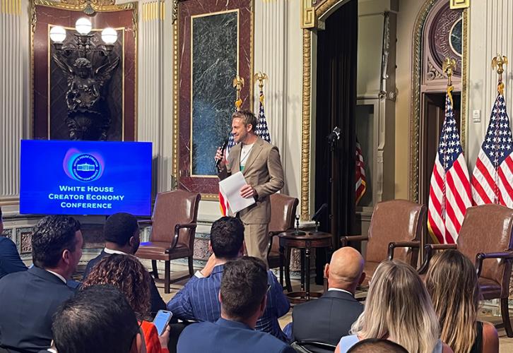 A wide shot of Janelle standing before a seated crowd holding a microphone in his right hand and papers in his left hand. To the left is a flatscreen with the White House logo and “White House Creator Economy Conference” in white. 