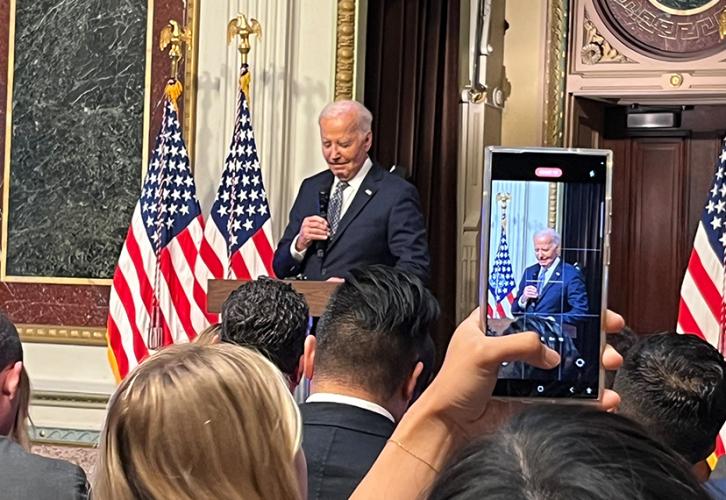 Biden stands behind a podium. His gaze is lowered; in his hand is a microphone. Members of the crowd stand in the photo’s foreground with their backs to the camera. One crowdgoer raises their arm to make a recording of Biden on their cellphone.