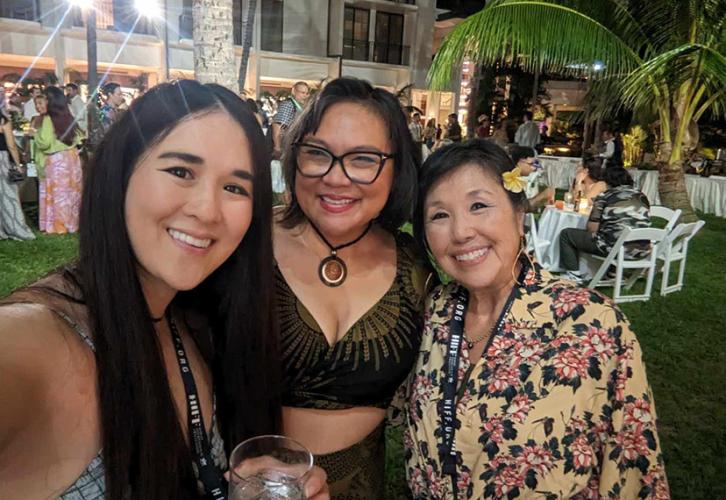 Three women pose for a selfie outdoors. People mingling at a party can be seen in the background. There is a palm tree on the right.