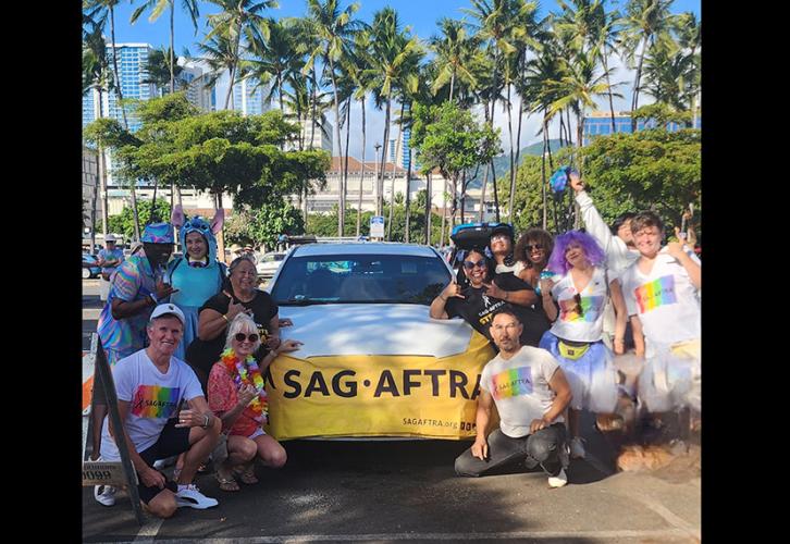 Members dressed in pride colors gather around a white car adorned with a yellow SAG-AFTRA banner. In the background are a group of palm trees and the city of Honolulu.