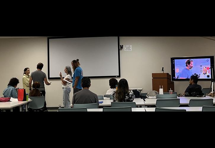 A group of diverse actors are in a classroom with a large TV screen on the right projecting two actors working on a scene together.