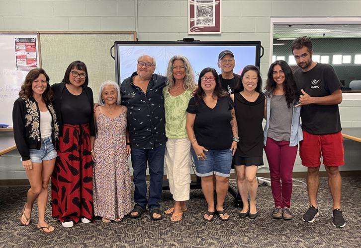 A diverse group of actors stand in a classroom in front of a large TV screen.