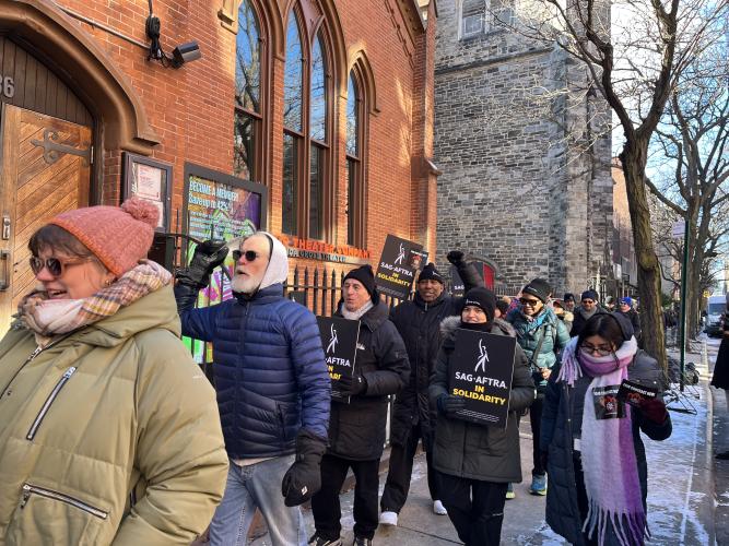 A group of people march past Atlantic Theater carrying black signs with “SAG-AFTRA in Solidarity” in white and yellow text.