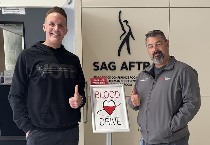 Two individuals stand on either side of a silver and white sign. There is a black SAG-AFTRA logo on the wall behind them.