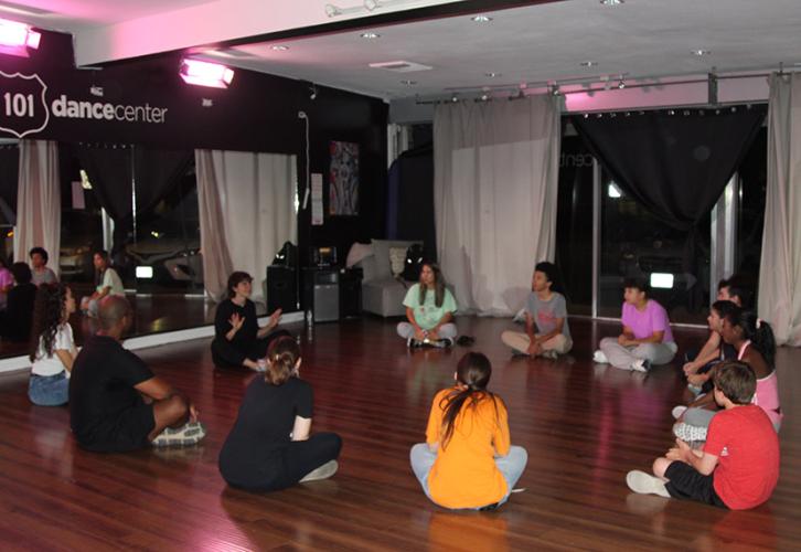 A group of individuals sit on the floor in a circle in a dance studio. 