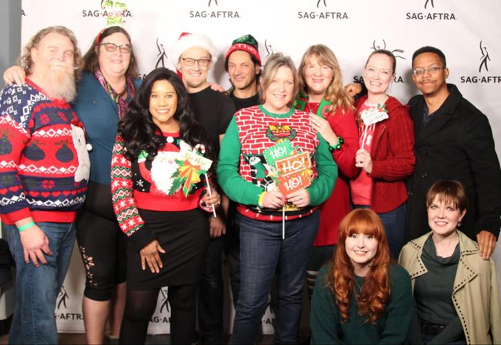 A group of individuals wearing holiday sweaters pose in front of a white backdrop with black SAG-AFTRA logos on it.