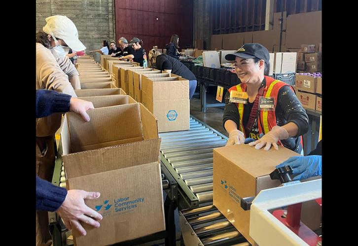 A group of individuals pack brown boxes on a conveyor belt in a warehouse. 