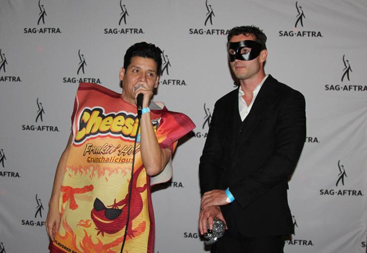 Two individuals in Halloween costumes stand with a mic in front of a step-and-repeat with SAG-AFTRA logo’s (black figure and its upraised right arm with “SAG-AFTRA” underneath) repeated on the white background.