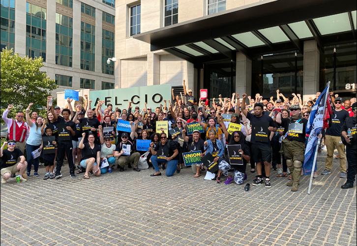 A large and diverse group of people stands outside a large city building. There is a large AFL-CIO sign behind the group. Many wear black “SAG-AFTRA Supports WGA” shirts and carry various signs that say, “Union Strong,” “SAG-AFTRA Strong” and “Solidarity.”