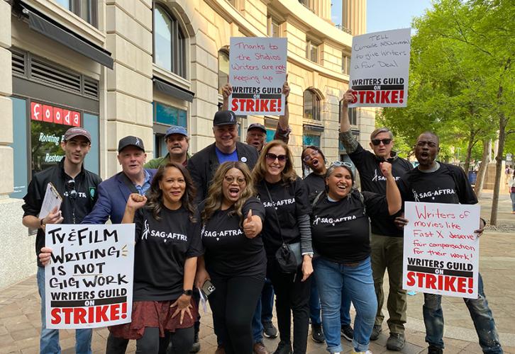 A diverse group of individuals stand outside on the pathway in front of a large building. Each person wears a black SAG-AFTRA T-shirt. Some people are cheering, others give a thumbs-up to the camera, while four people hold WGA picket signs. 