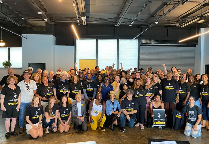 A large and diverse crowd of people stand in a large open room. Many of the people are wearing black “SAG-AFTRA Strong” T-shirts. Many of the people are smiling. A woman in the front holds a black and yellow “SAG-AFTRA on Strike” picket sign.