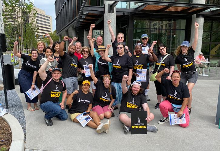 A large and diverse group of people on the sidewalk outside of a building. They all wear black SAG-AFTRA T-shirts. They all have their fists in the air. A man sitting in the front row holds a black “SAG-AFTRA Strong” picket sign, while others hold informational pamphlets.