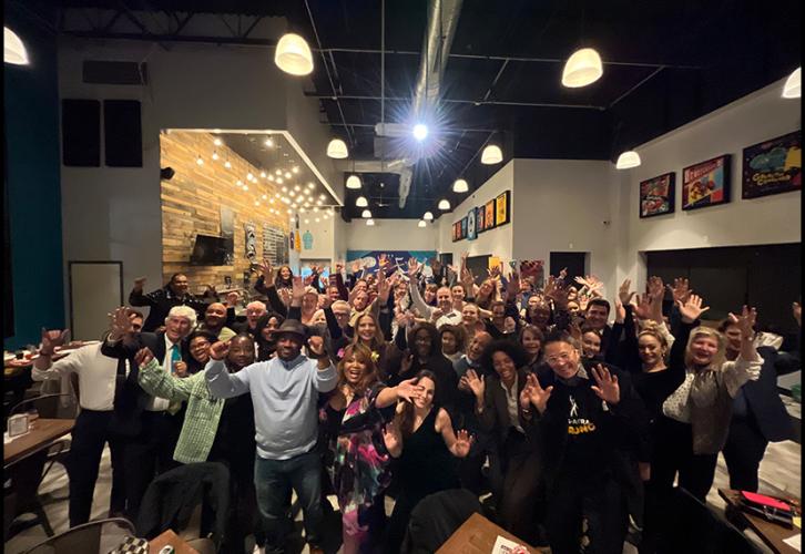 A group photo of multiple, diverse individuals in a restaurant. Attire ranges from business casual to formal. All in the foreground have their arms raised in a cheer.