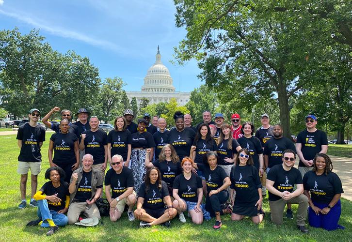 A diverse group of individuals gather in a park with the United States Capitol building behind them. The people are in two rows: The back row is standing, and the front row is kneeling. The individuals are wearing black “SAG-AFTRA Strong” T-shirts. Everyone is smiling, and some members have a raised fist.