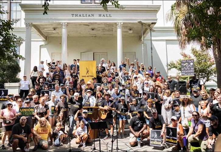 A diverse group of people stand in front of a large, white government building. Many hold various strike signs, including handmade signs. In the back rows of the group is a yellow SAG-AFTRA banner. 