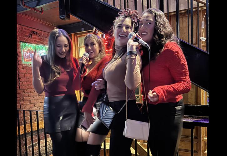 Four SAG-AFTRA New England Local members wearing red for the holiday party sing karaoke on the Wild Rover Bar stage in front of a large audience of members.