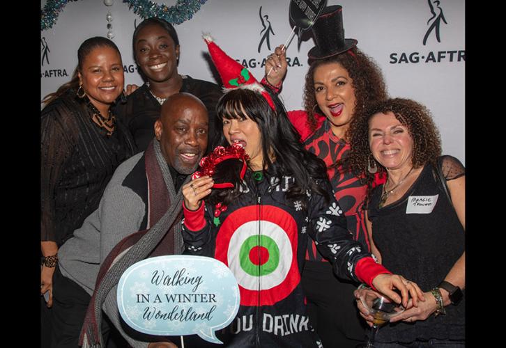 A diverse group of seven SAG-AFTRA New England members pose in front of the SAG-AFTRA step-and-repeat, some holding holiday props and wearing festive hats.