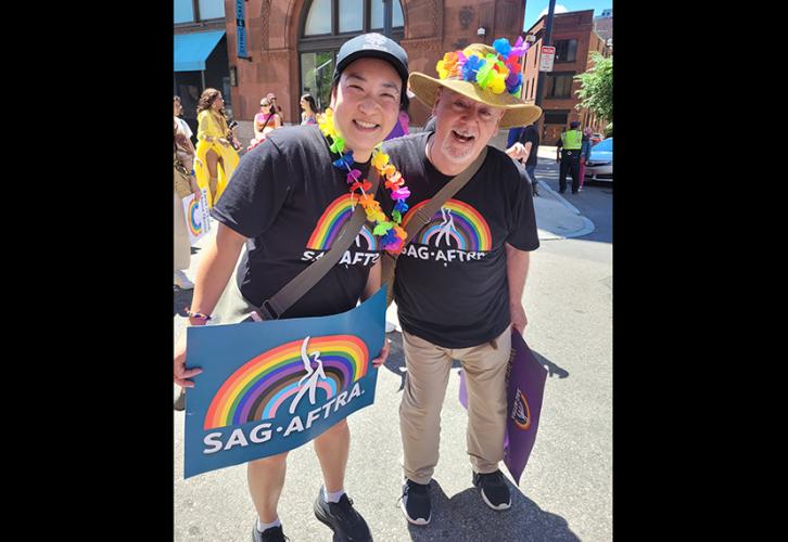 IATSE Local 481 President Kimmie Johnson wearing an IATSE baseball cap, a lei and a SAG-AFTRA Pride shirt poses with a man wearing a straw hat, a lei and a SAG-AFTRA Pride shirt as they walk down Berkeley Street in the Boston Pride Parade. 