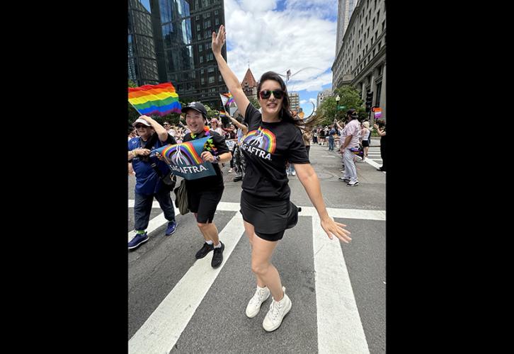 New England Local member Megan Carroll wears a SAG-AFTRA pride T-shirt and holds her right arm up during the pride parade in Boston. A woman on her right holds a SAG-AFTRA rainbow sign. The John Hancock Tower and marchers holding various pride flags can be seen in the background.