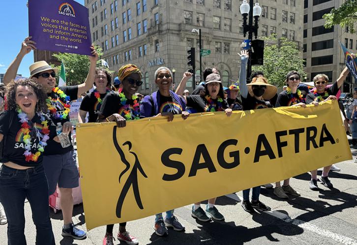 A diverse group of people wearing SAG-AFTRA pride T-shirts hold a large gold horizontal banner that reads “SAG-AFTRA” while walking down Berkeley Street in the Boston Pride for the People Parade. A woman in the middle wearing a baseball cap holds the banner with her left hand while raising her right hand in a solidary fist.  Additional people in the parade hold pride posters and flags. 