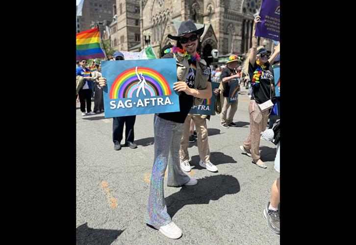 Union member Brian Bates, wearing sequined pants, a SAG-AFTRA Pride T-shirt, a tan vest and a black cowboy hat and holding a SAG-AFTRA Pride sign, walks in the Pride parade on Boylston Street with the Old South Church and pride flags in the background.