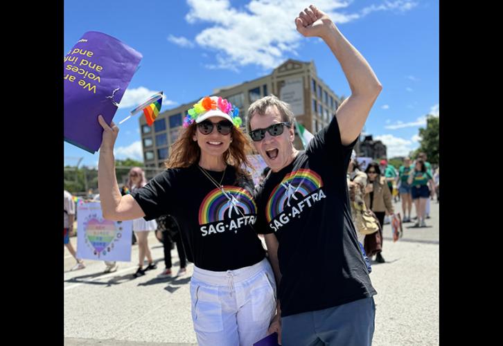 A woman wearing a SAG-AFTRA pride T-shirt and a white baseball cap with a lei sitting on the brim of the cap holds up a pride sign in her right hand. A man in a SAG-AFTRA pride T-shirt raises his left arm with his hand in a solidary fist.  