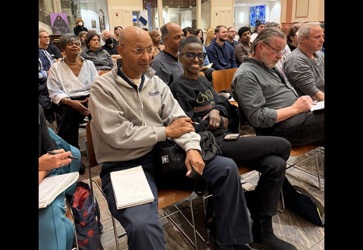 Christoper James in blue pants and gray top and Gilda James in all black sit in the front row of a diverse group of people sitting listening to the conservatory.