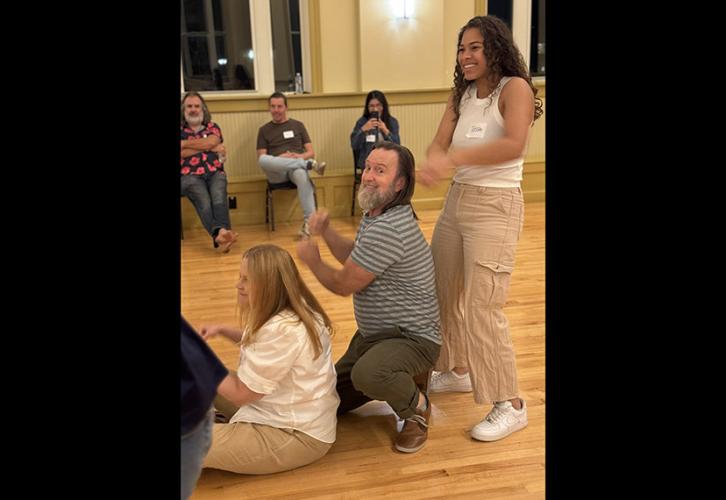 A woman makes rowing movements while turning perpendicular to the camera and sitting on the floor. A man crouches on the ground behind her, making similar motions and smiling at the camera. Another woman stands behind them, smiling at something off-camera. Three members sit in chairs in the background, watching the exercise.