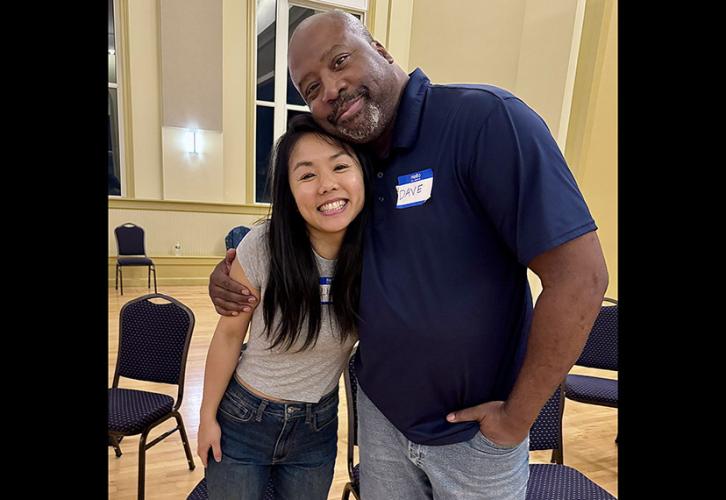 A man and a woman wearing jeans and short sleeves, stand side by side with one arm on each other’s shoulder.