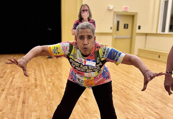 A woman in black pants and a colorful top blows air from her mouth and stretches her arms outward.