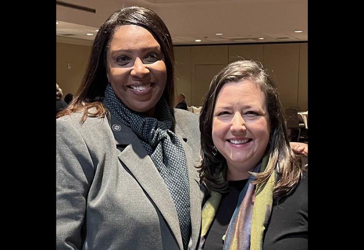 A woman in a gray blazer and a woman in a black top and colorful scarf smile for the camera. 